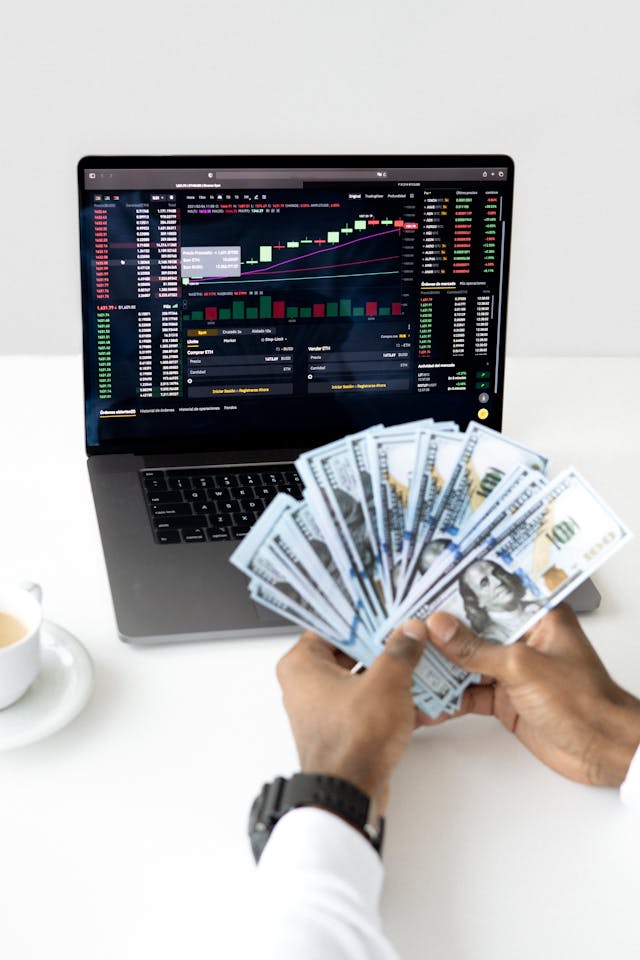 Person holding a fan of hundred-dollar bills in front of a laptop displaying financial charts, symbolizing proactive capital planning.