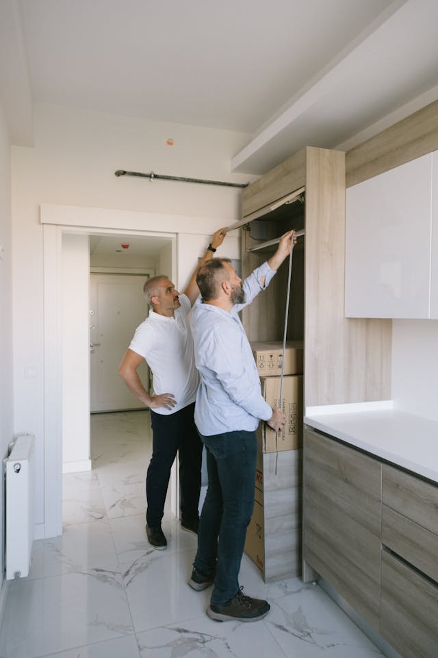 Two workers measuring cabinetry and inspecting interior fixtures, representing planning and assessment of building systems.